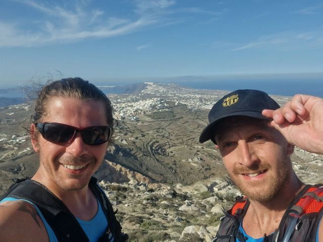 Two runners in sunglasses taking a selfie above Santorini's white villages and the sea.