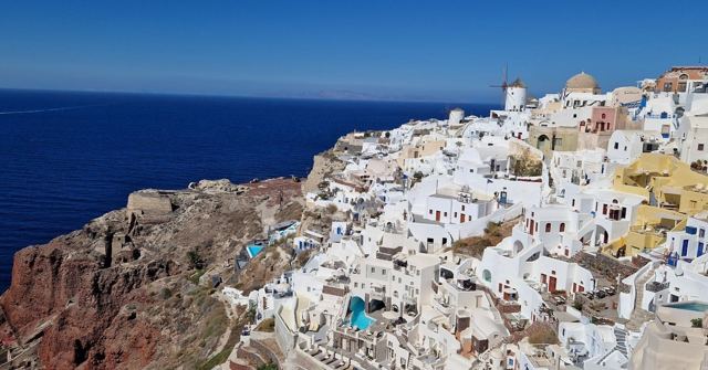 White houses and a windmill clinging to a cliff above a deep blue sea.