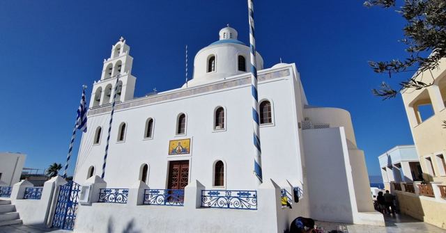 A white Greek Orthodox church with a bell tower against a deep blue sky.