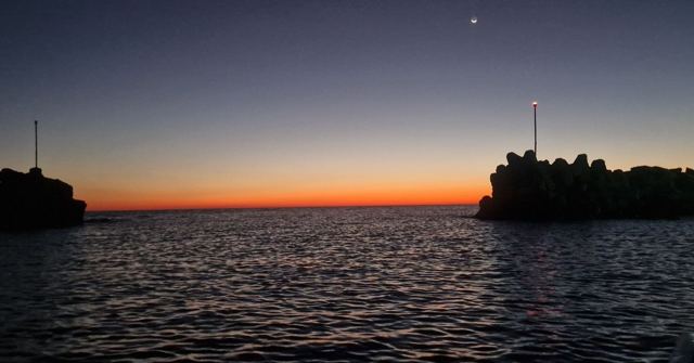 Dark silhouetted rocks and a small beacon against a deep orange and blue twilight sky over the sea.