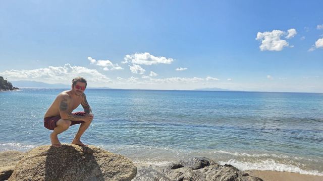 A man crouching on a rock at the edge of a calm, clear blue sea.
