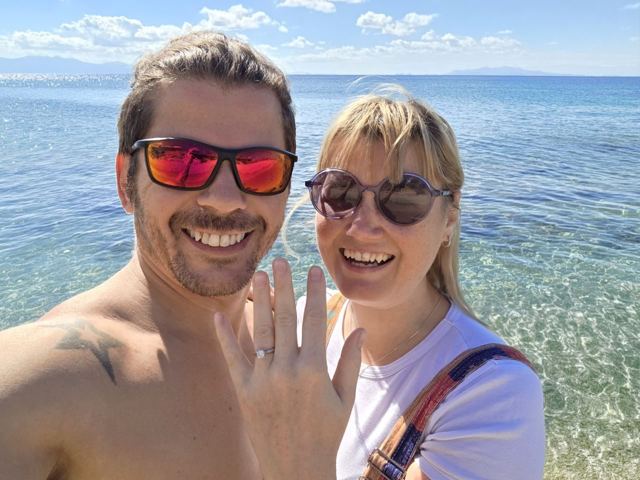 A smiling man and a woman taking a selfie in front of a turquoise sea.