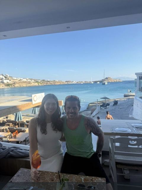 A man and a woman standing on a seaside terrace with white buildings and a bay behind.