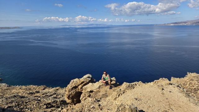 View across a deep blue sea from sandy rocks, with a figure sitting on the rocks.