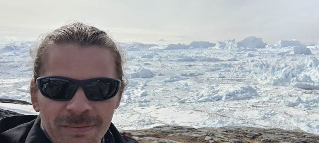 A man in sunglasses taking a selfie in front of a fjord filled with huge icebergs.
