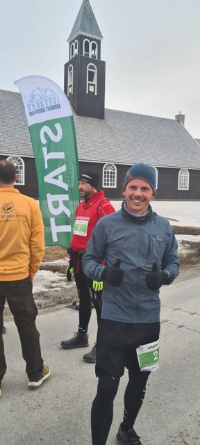 A smiling man in a jacket and beanie giving thumbs up at a marathon start in Ilulissat.