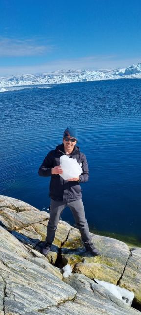 A man in a beanie holding a lump of clear ice by deep blue water with icebergs behind.