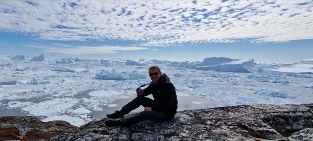 A man sitting on rocks above a fjord crowded with white icebergs under a dappled sky.