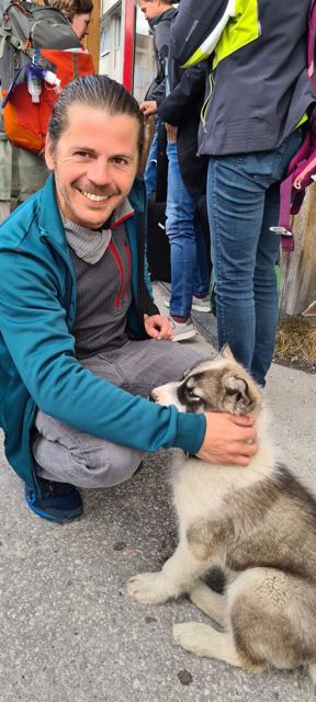 A smiling man crouching to stroke a fluffy grey sled dog puppy.