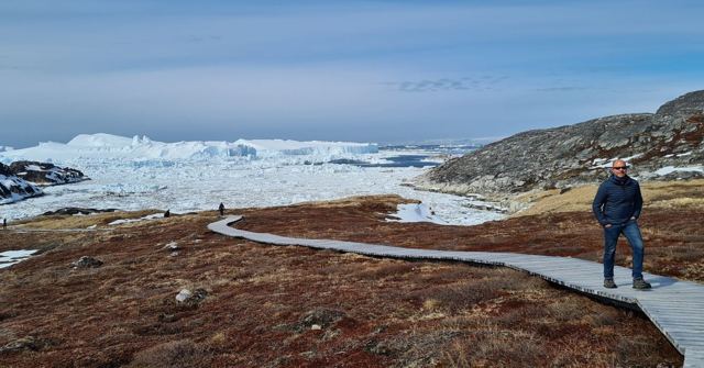 A man standing on a boardwalk across brown tundra, with a fjord packed with ice behind.