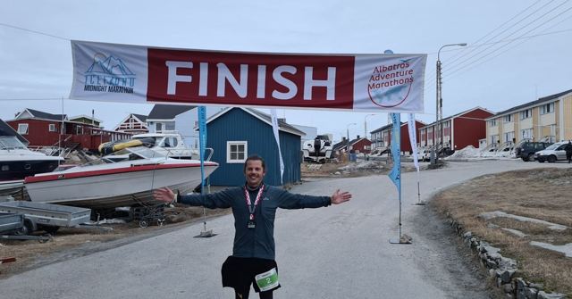 A man in running kit with a medal, arms outstretched under a red FINISH banner on a street.
