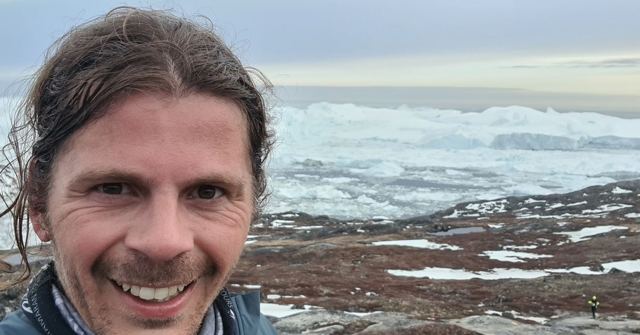 A smiling man taking a selfie with a fjord full of icebergs and tundra behind him.