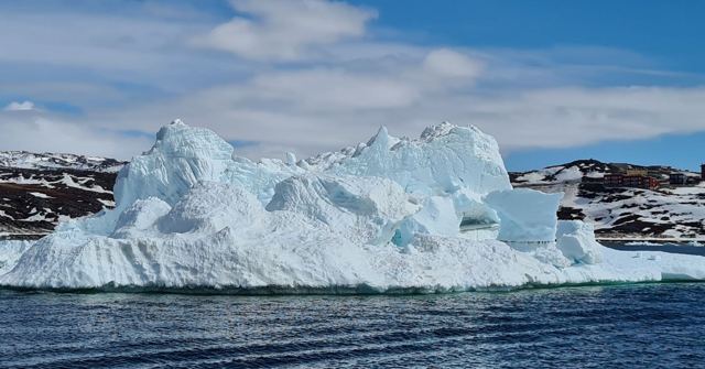 A large white iceberg floating on dark blue sea under a bright blue sky with clouds.
