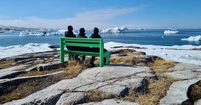 Three people seen from behind on a green bench, looking over rocks and a sea dotted with icebergs.