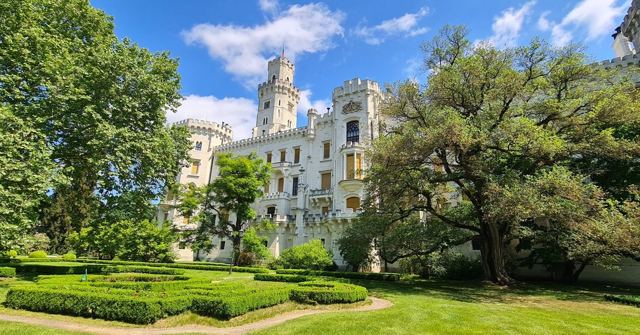A white neo-Gothic castle with turrets behind manicured hedges and large green trees.