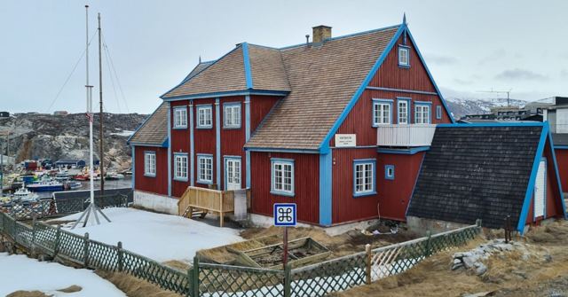 A red two-storey wooden house with blue trim and a picket fence beside snow.