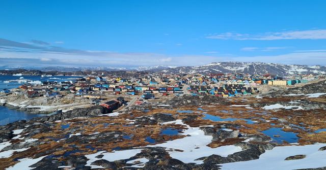 A town of colourful houses across rocky ground with patches of snow and the sea behind.
