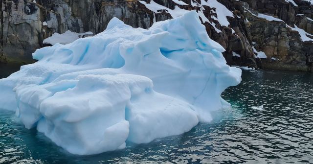 A bright blue and white iceberg floating in dark water beside rocky cliffs.