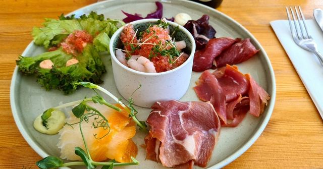 A plate with a bowl of prawns, cured meat slices, salad and smoked fish on a wooden table.