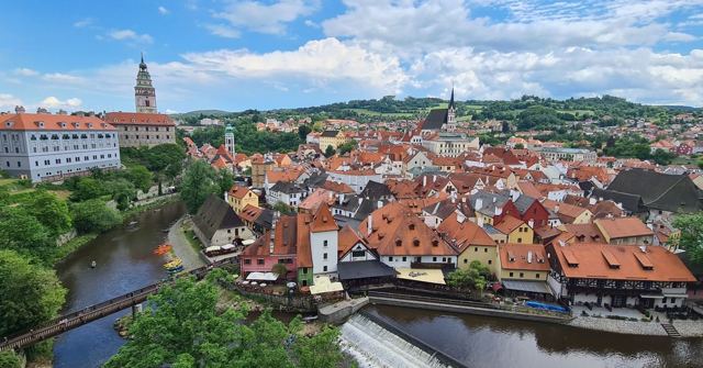 A panoramic view of a town of red-roofed houses on a river bend, with a castle tower.