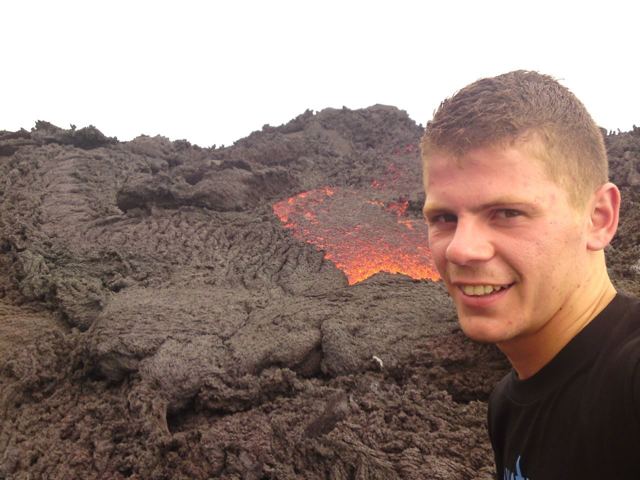 A man smiling in front of glowing orange lava flowing between black rocks on Pacaya volcano.