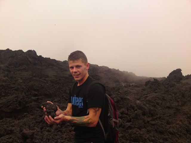 A man crouching on black lava fields in the mist, holding a piece of volcanic rock.