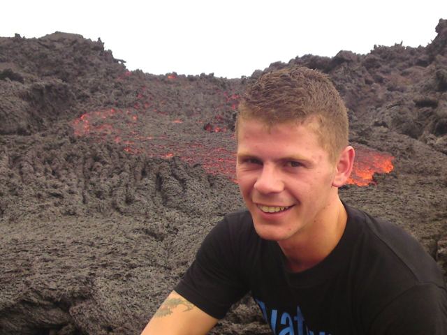 Close-up of a man smiling with a stream of red lava visible behind him on Pacaya volcano.