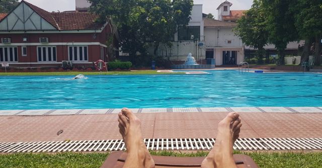 A pair of bare feet in the foreground of a swimming pool with a red-brick building behind.