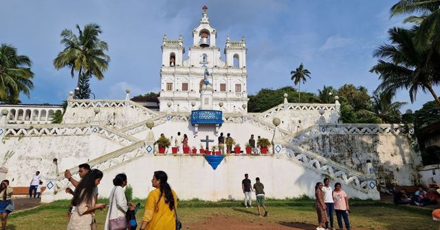 A brilliant white church with a tall bell tower and zig-zag staircase, palm trees and visitors in front.