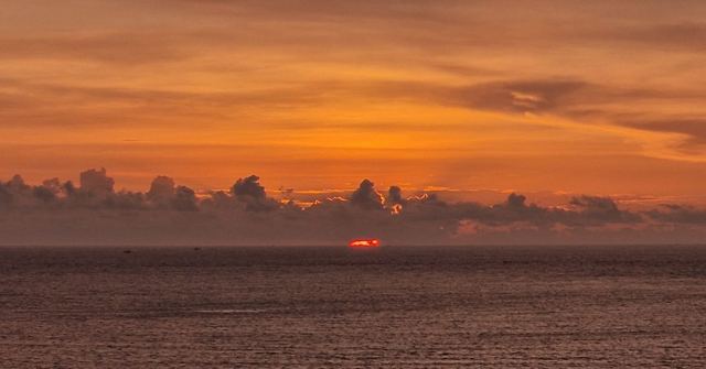 A deep orange sunset sky over a dark, calm sea with the sun just touching the horizon.