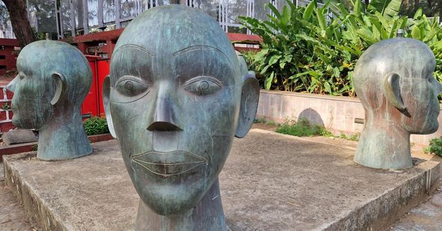 Large bronze sculptures of human heads arranged in a garden with plants behind.