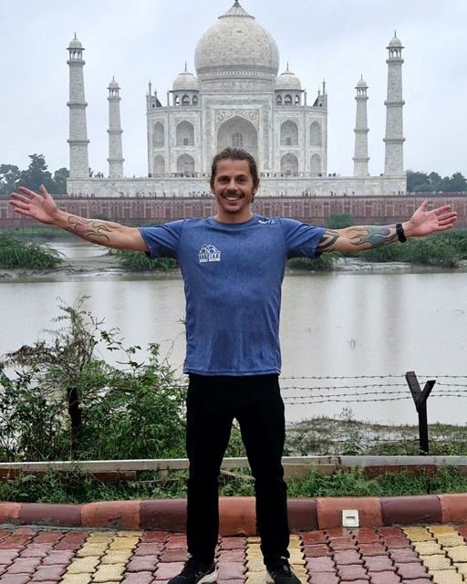 A smiling man with arms outstretched in front of the Taj Mahal across a river.