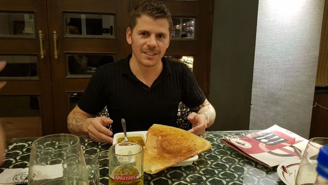 A smiling man at a restaurant table with a huge rolled dosa in front of him.