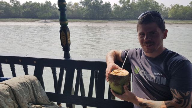 A man holding a coconut with a straw on a boat on a wide river lined with mangroves.