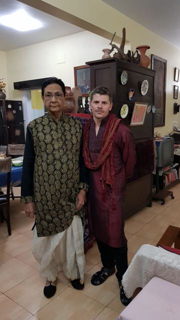 A man in a red kurta standing beside an older man in traditional Indian dress in a living room.