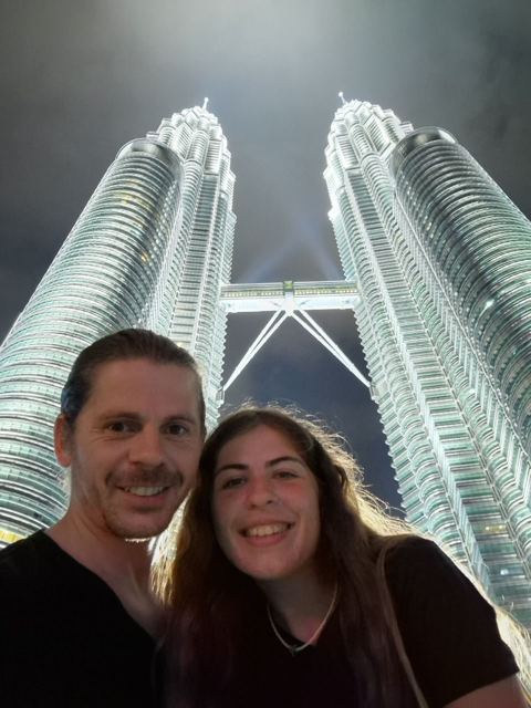 A man and a woman taking a selfie below the illuminated Petronas Towers.