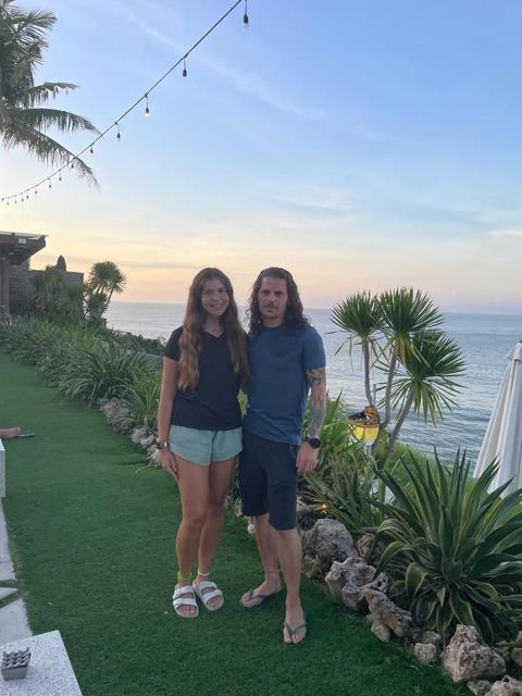 A man and a woman standing on a clifftop lawn by the sea at sunset.