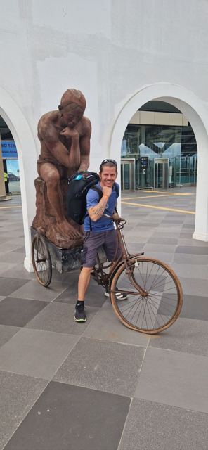 A man leaning on an old bicycle beside a large bronze sculpture outside a terminal.