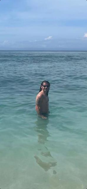 A man with long hair standing waist-deep in turquoise sea.