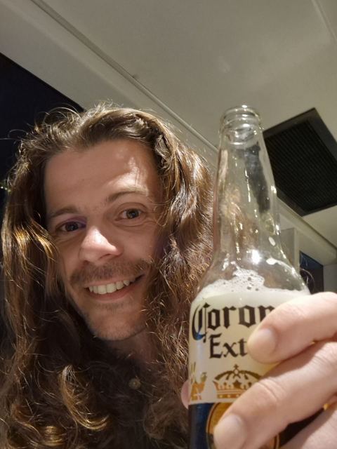 A man with long hair smiling and holding up a bottle of Corona.