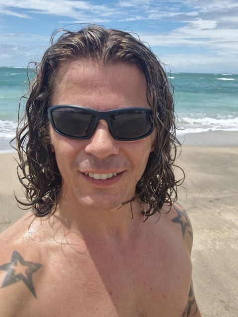 A smiling man in sunglasses with wet hair on a beach with waves behind.