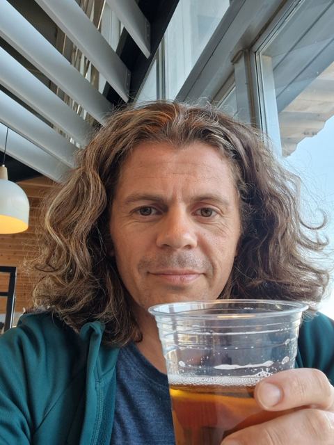 A man with long hair raising a plastic cup of beer at an airport.
