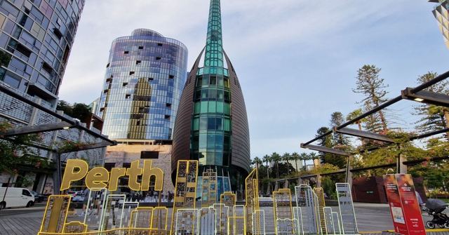 Large yellow letters spelling Perth in front of a glass spire tower and modern buildings.