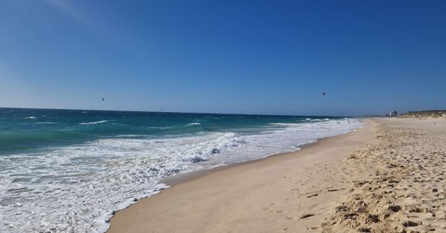 A wide white sand beach with waves breaking under a clear blue sky.