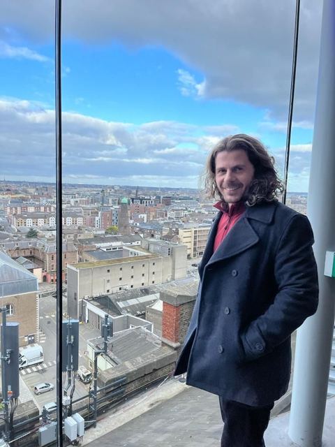 A man in a dark coat standing by the glass wall of a bar with Dublin rooftops below.
