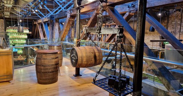 Whiskey barrels and display cases inside a distillery with exposed iron beams and a glass walkway.