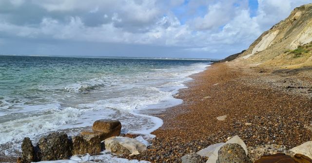 Waves breaking on a pebble beach below sandy cliffs under a cloudy blue sky.