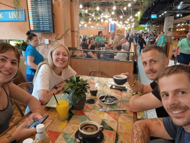 A group of friends smiling around a table in a busy airport restaurant.