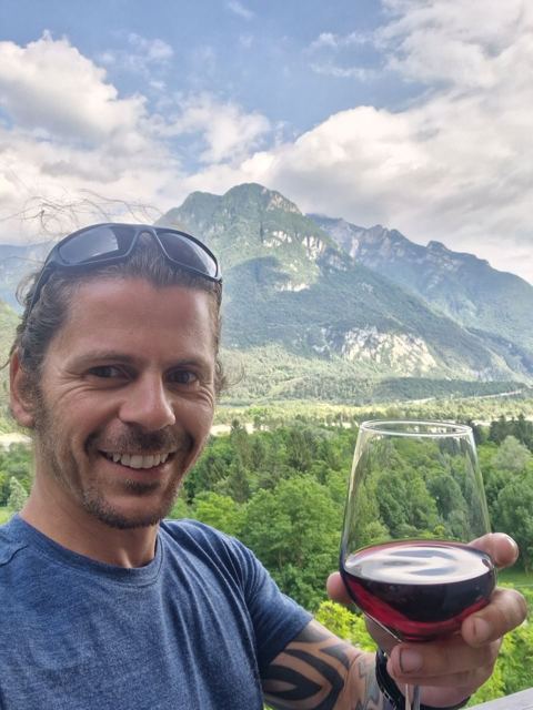 A smiling man raising a glass of red wine in front of green mountains.