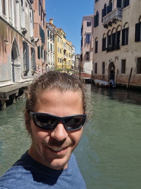 A man in sunglasses taking a selfie beside a canal lined with old buildings in Venice.